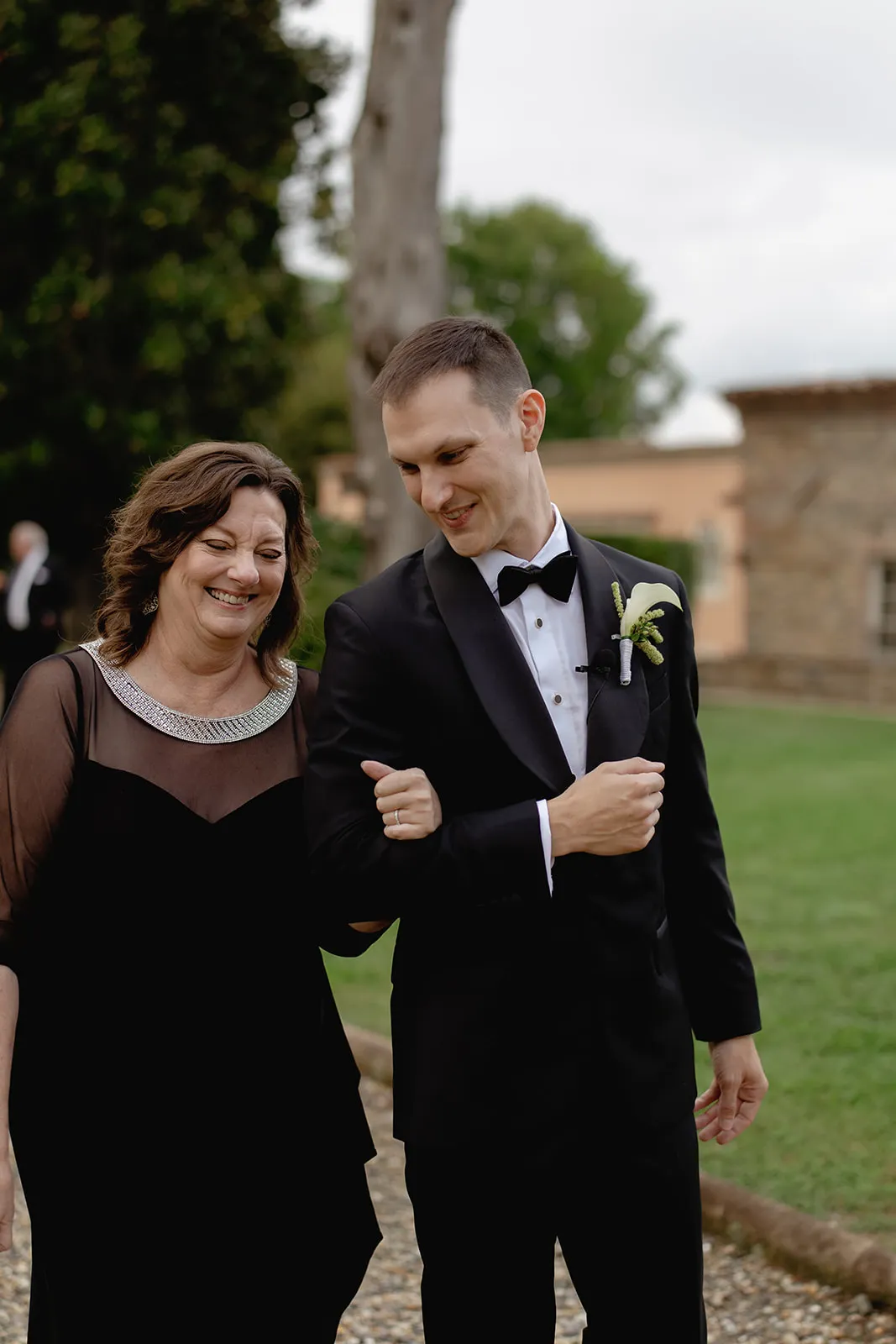 A smiling woman in a black dress with sheer sleeves walks arm-in-arm with a man in a black tuxedo and bow tie. They are outdoors on a stone path with grass and trees in the background. The man wears a white boutonniere. Both look happy. Tuscan Wedding Photographer