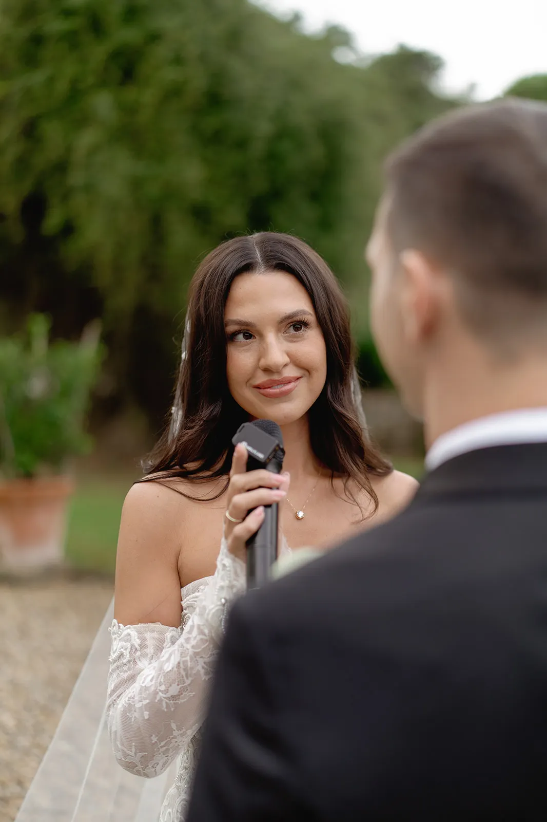 A bride in an off-the-shoulder white dress holds a microphone, smiling warmly at the groom, who is facing her in a black suit. They stand outdoors with greenery and a potted plant blurred in the background, creating a romantic atmosphere. Tuscan Wedding Photographer