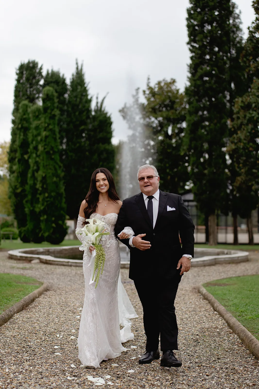 A bride in a white, off-shoulder lace wedding dress holds a bouquet of white flowers, smiling as she walks arm-in-arm with an older man in a black suit. They walk on a gravel path in front of tall trees and a fountain in a garden. Tuscan Wedding Photographer
