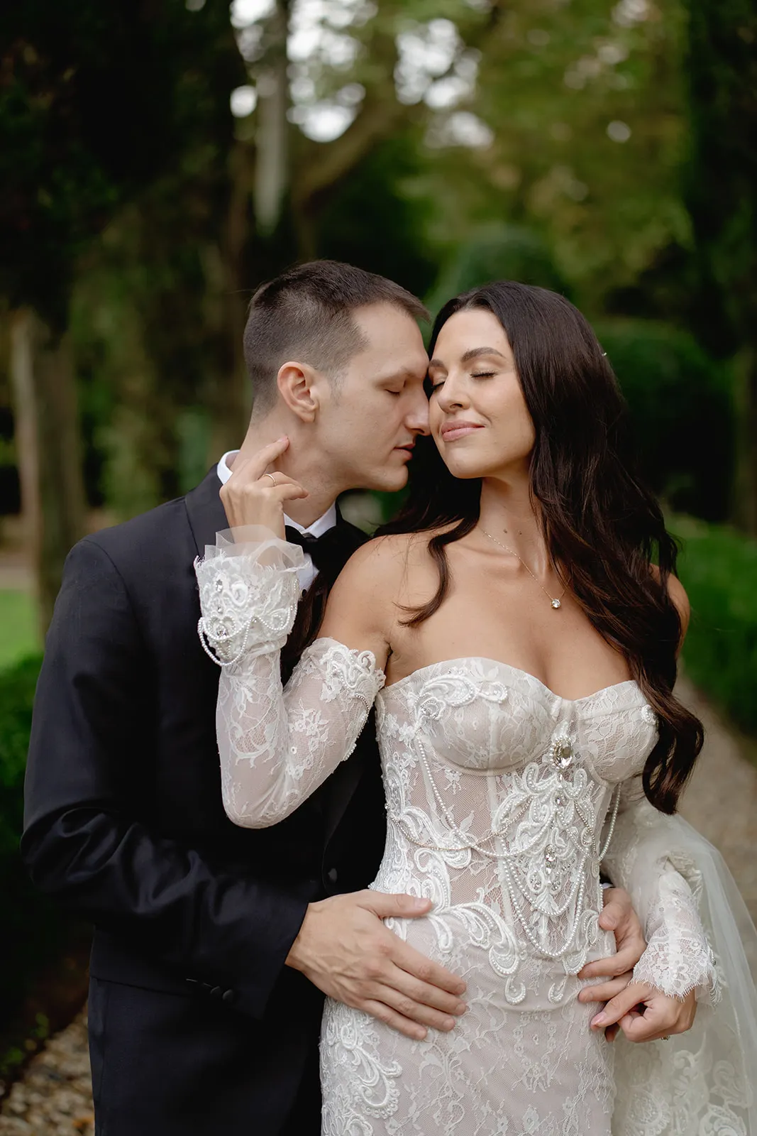 A groom in a black tuxedo gently embraces a bride in an off-the-shoulder lace wedding dress. The bride closes her eyes, smiling softly, while the groom lovingly kisses her temple. They stand on a tree-lined path surrounded by greenery. Tuscan Wedding Photographer