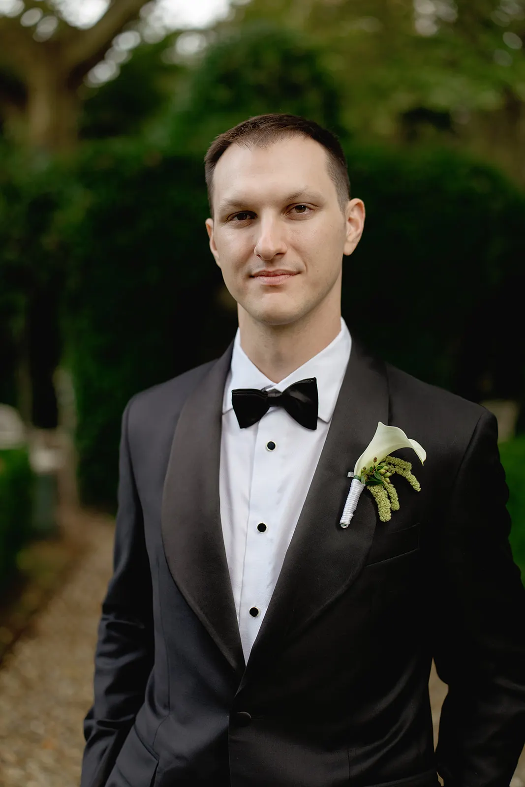 A man stands outdoors wearing a black tuxedo with a white shirt, black bow tie, and a white calla lily boutonniere. He has short brown hair and a slight smile. The background shows green foliage and a blurred path, suggesting a garden setting. Tuscan Wedding Photographer