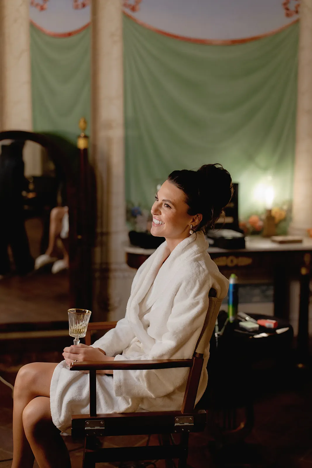A woman in a white robe sits on a chair, smiling and holding a glass with a patterned design. Her dark hair is in a loose updo. The room behind her is softly lit, with green draped fabric, a mirror, and elegant, vintage-style decor. Tuscan Wedding Photographer