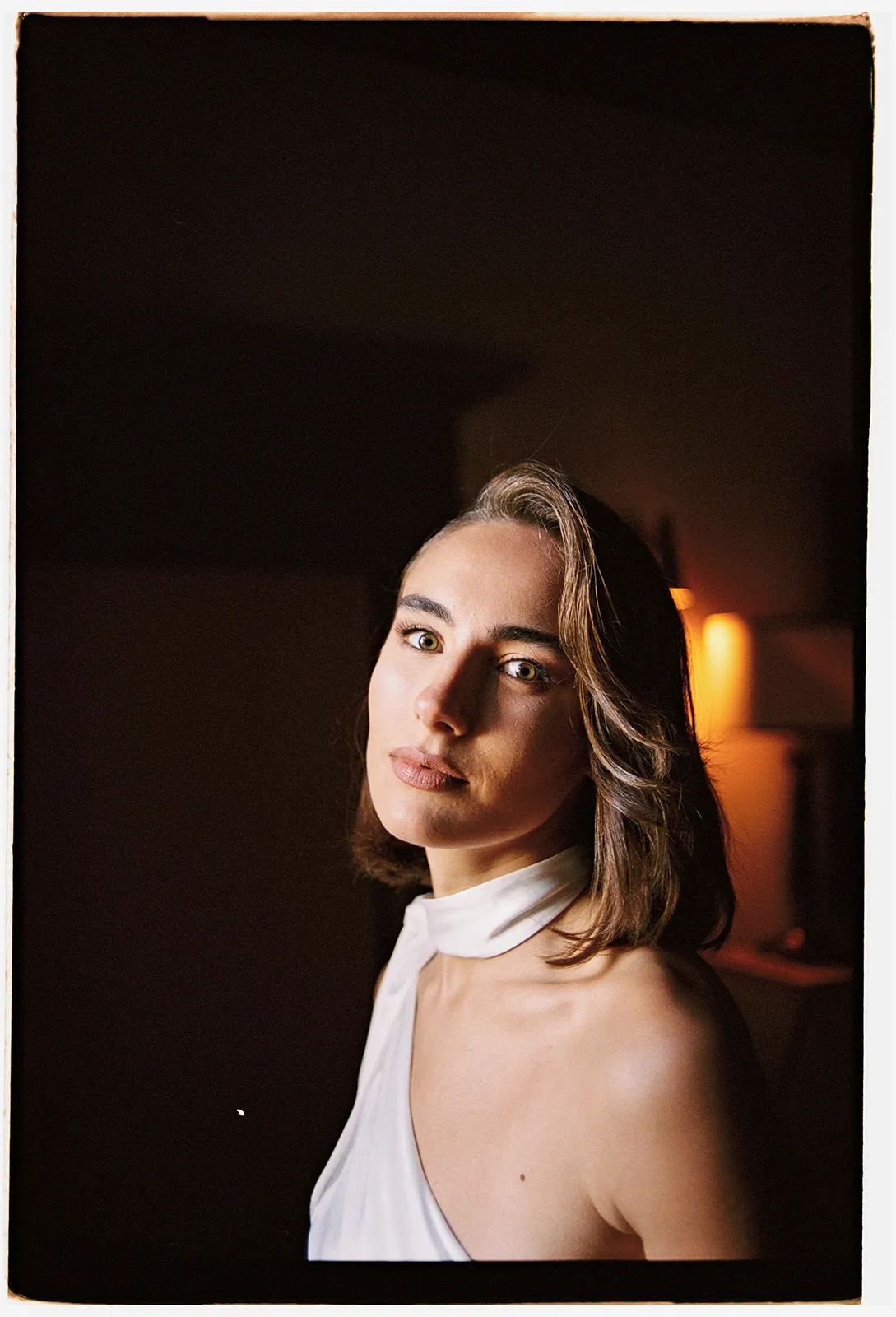 A woman with straight brown hair and light skin wears a white, one-shoulder top and scarf. She looks calmly at the camera in a dimly lit room with a warm light in the background, highlighting her face and shoulders against a dark backdrop—an image evocative of timeless elegance, perfect inspiration for engagement photos captured by a Tuscany wedding photographer. Italy and Tuscan Wedding Photographer