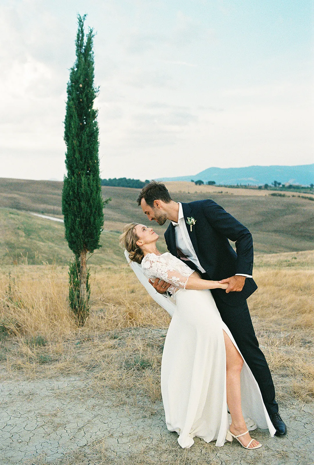 A groom in a black suit gently dips a smiling bride in a white gown outdoors, their joy radiating as if captured for engagement photos. They stand on dry grass in a field with rolling hills, a single tall cypress tree, and distant mountains under a lightly clouded sky—the perfect scene for any Tuscany wedding photographer to immortalize. Italy and Tuscan Wedding Photographer