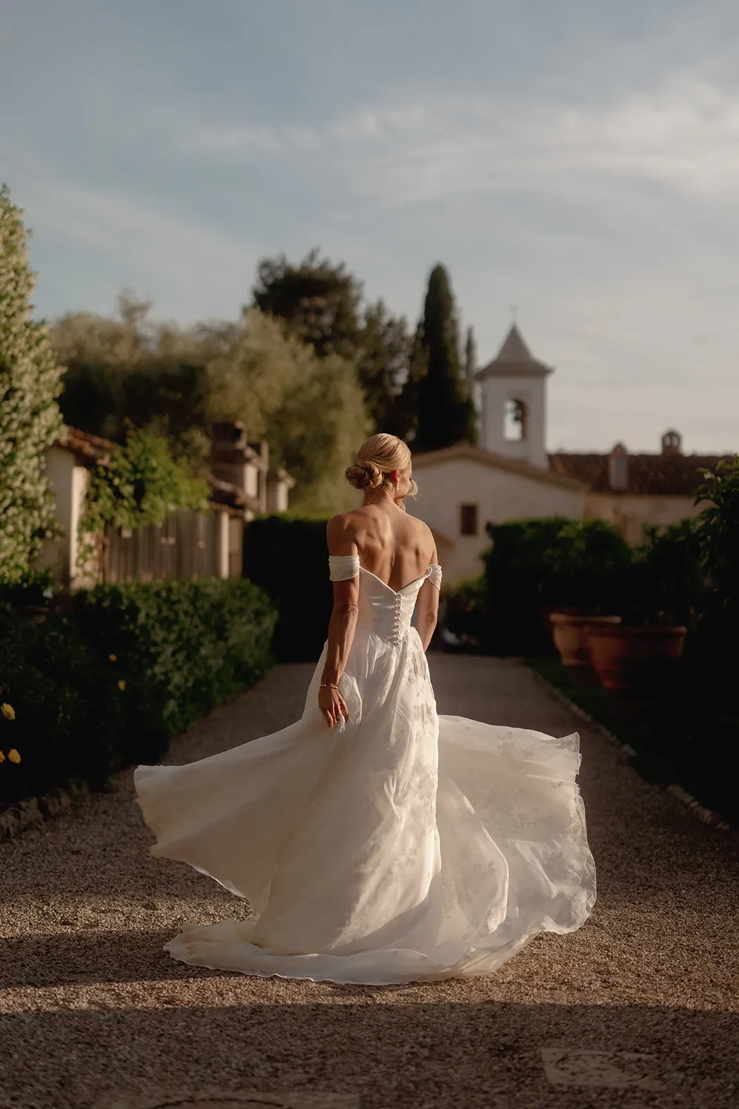 A woman in a flowing white wedding dress walks away on a gravel path lined with greenery, her back to the camera. Her blond hair is in a low bun. A sunlit church with a bell tower is in the background under a clear sky, capturing the enchanting atmosphere of a Destination Wedding in Italy. Italy and Tuscan Wedding Photographer