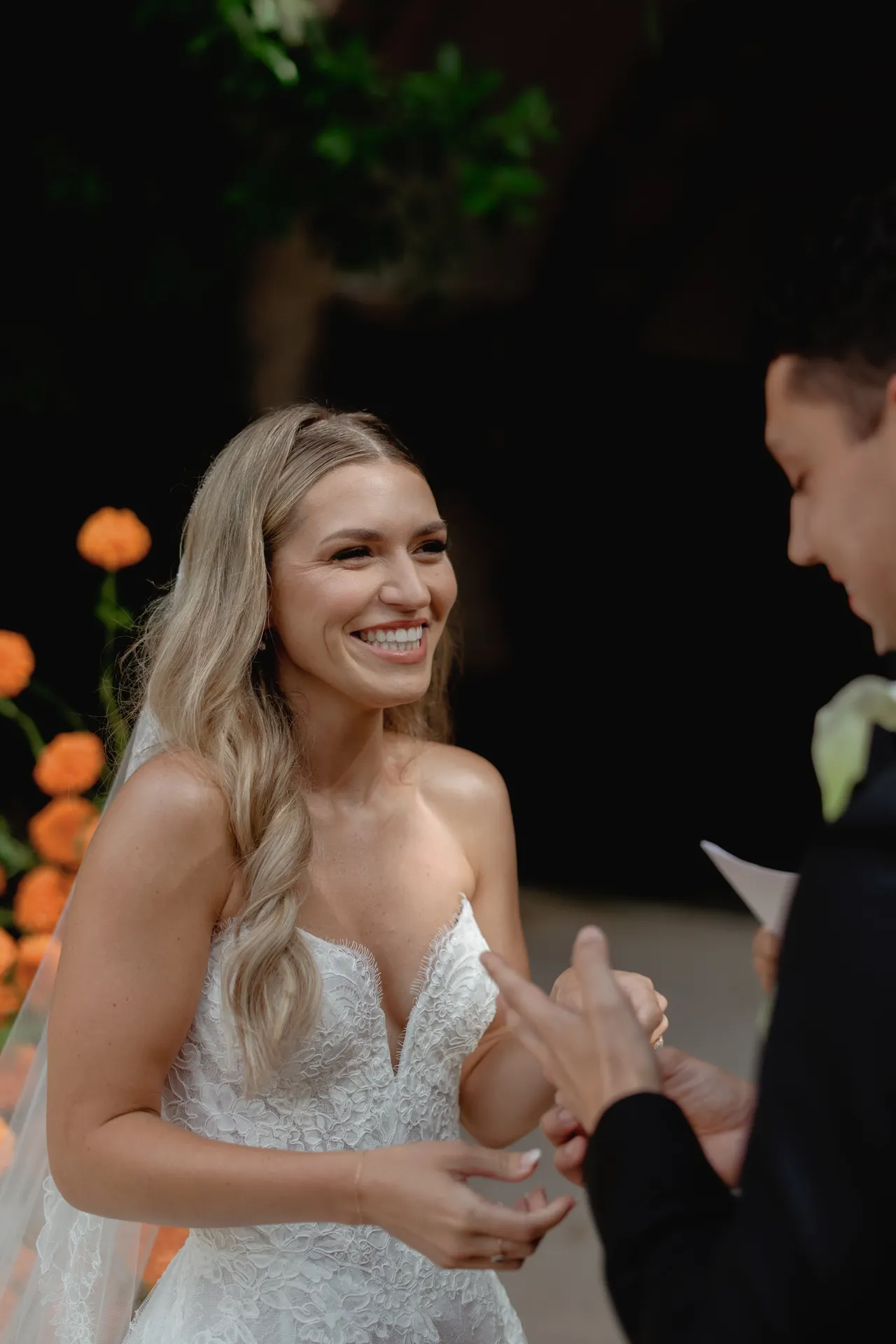 A smiling bride in a white lace dress stands facing someone placing a ring on her finger. She has long, wavy blonde hair and is outdoors near orange flowers, with greenery and a dark background behind her—capturing the romance of a Destination Wedding in Italy. Italy and Tuscan Wedding Photographer