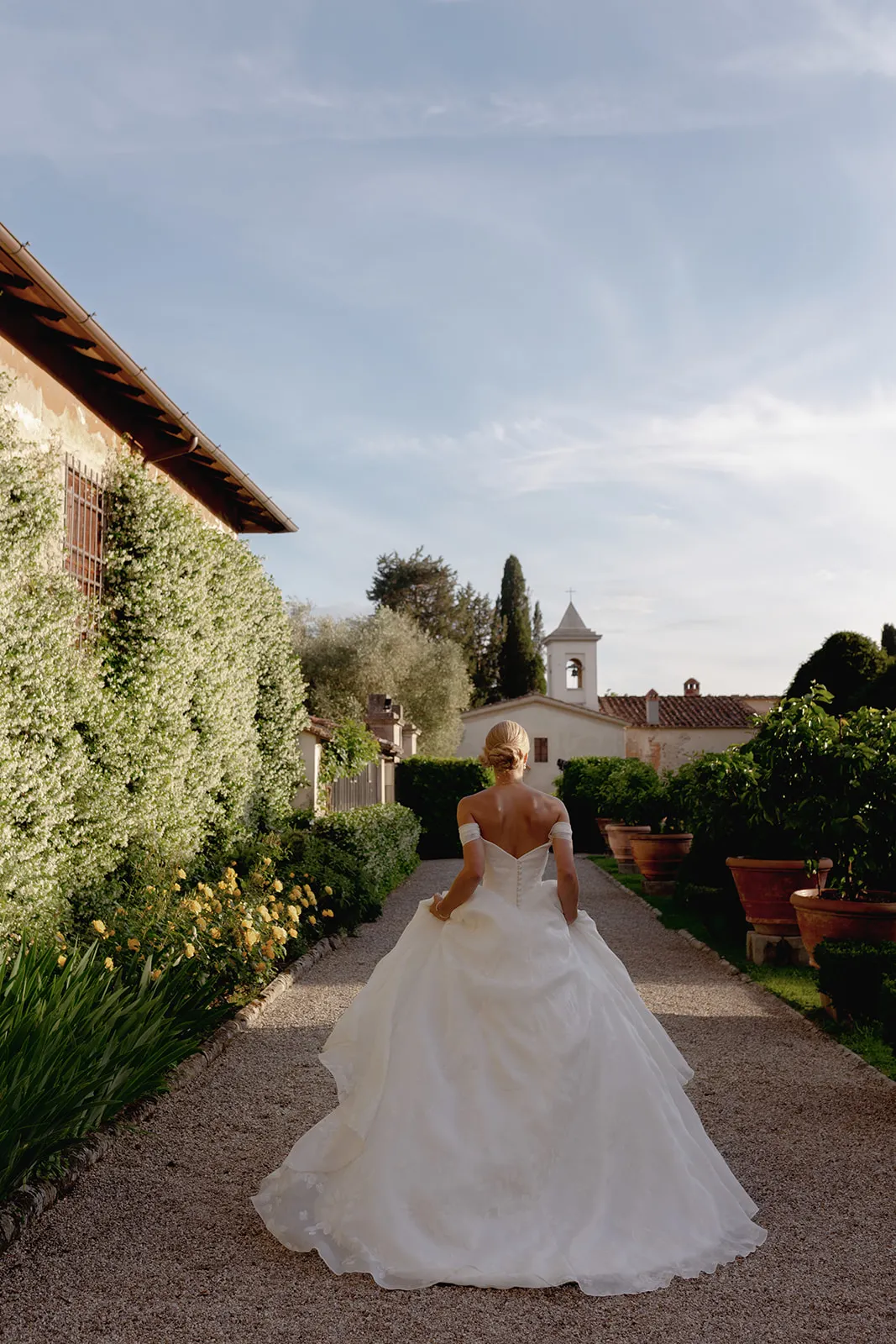 A bride in a white gown walks alone on a gravel path between lush green hedges and large potted plants, the serene setting epitomizing a Destination Wedding in Italy, with a bell tower and rooftops visible in the background under a partly cloudy blue sky. Italy and Tuscan Wedding Photographer