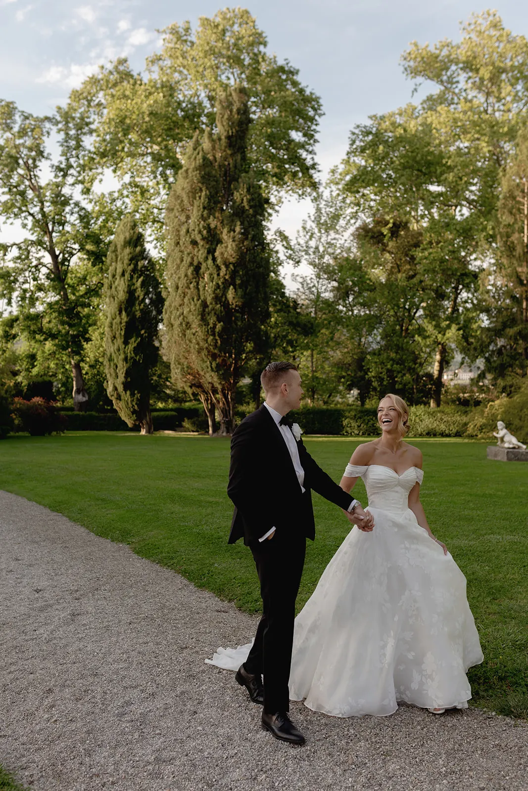 A bride in a white gown and groom in a black tuxedo walk hand in hand on a gravel path in a lush green park, smiling at each other, with tall trees and a statue in the background under a clear sky—a picturesque scene from their enchanting destination wedding in Italy. Italy and Tuscan Wedding Photographer