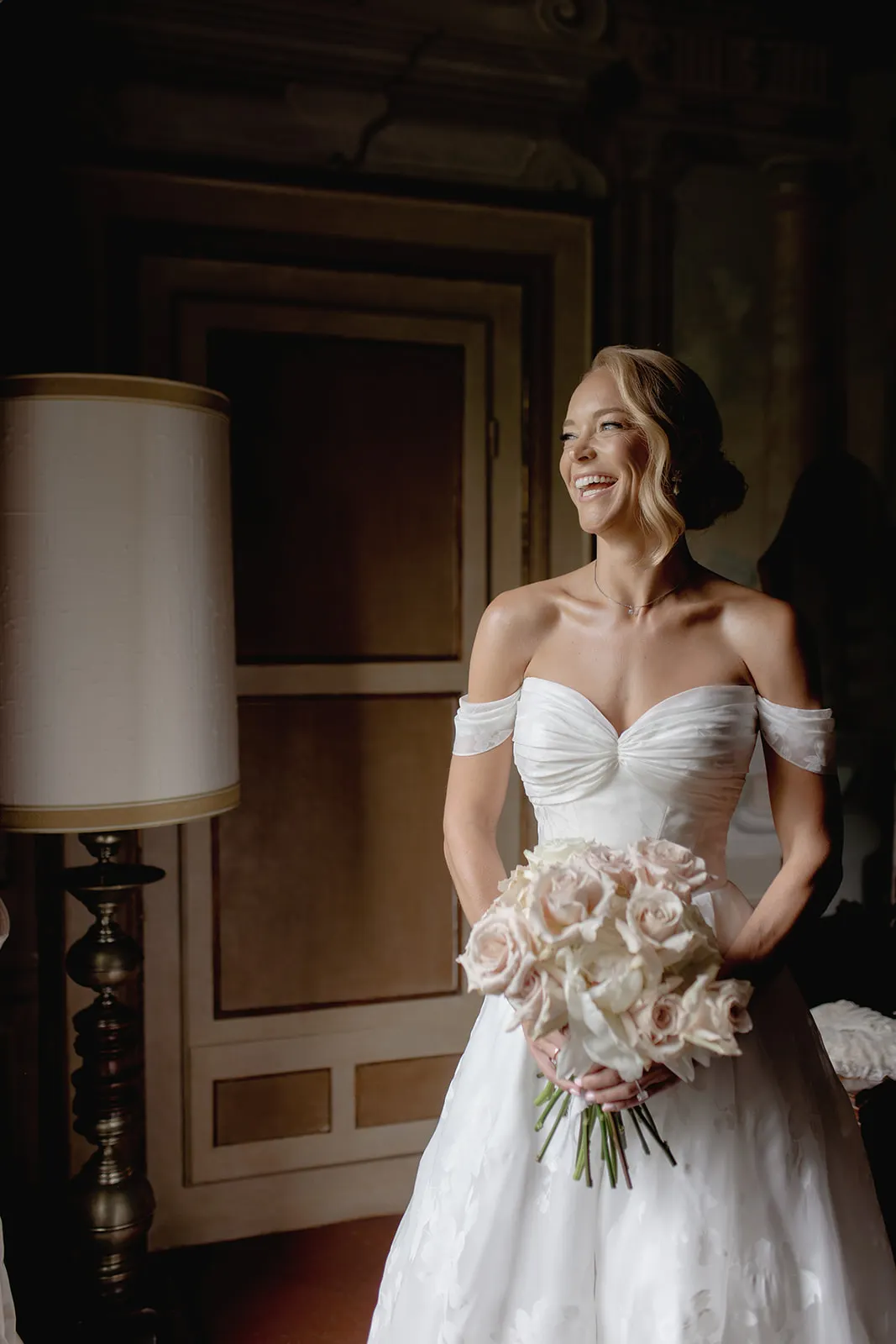 A smiling bride in an off-the-shoulder white gown holds a bouquet of pale pink and white roses. She stands indoors beside a tall lamp, with a closed door and ornate wall in the softly lit background—capturing the elegance and romance of a Destination Wedding in Italy. Italy and Tuscan Wedding Photographer