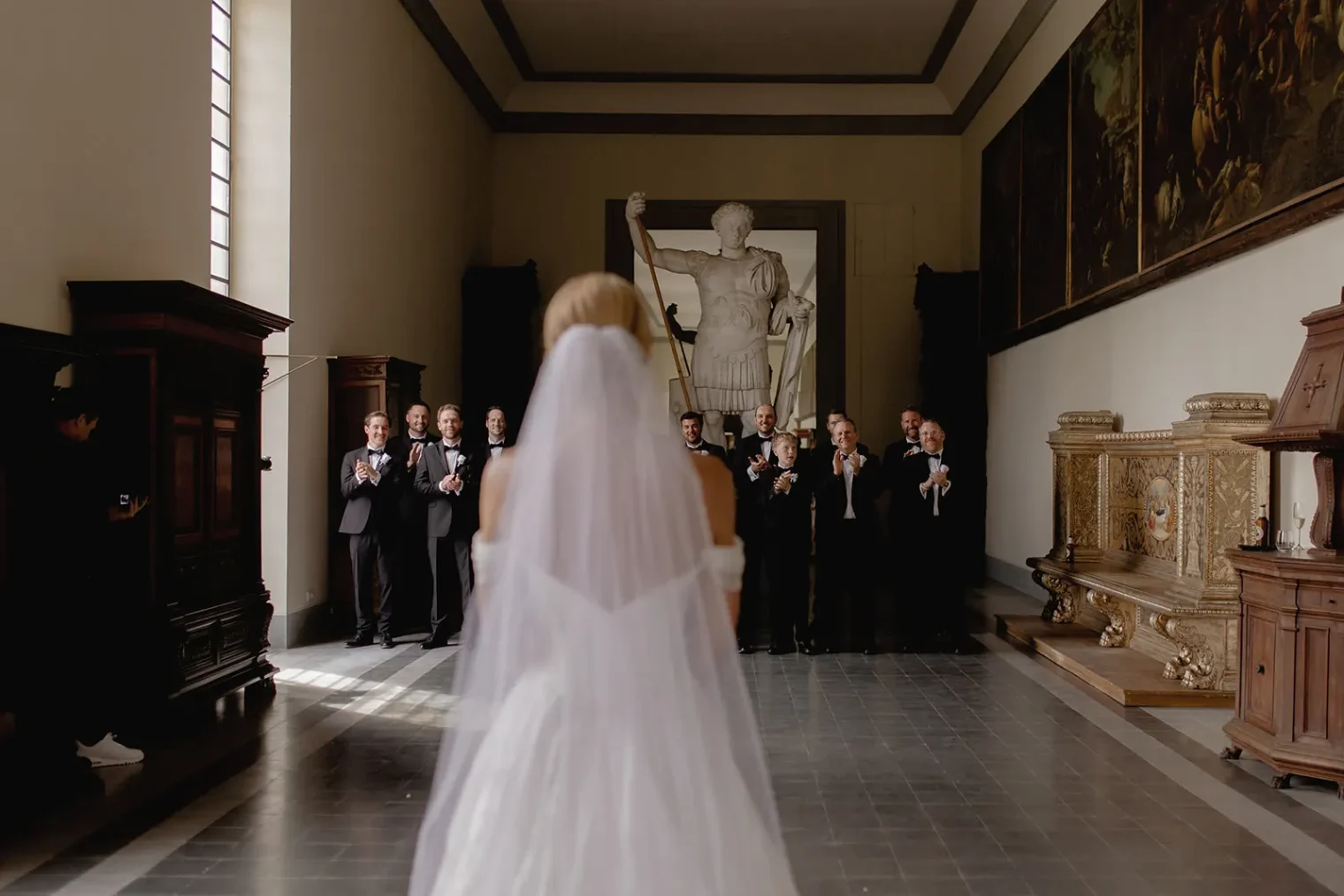 A bride in a white gown and veil stands in a grand hall facing a group of groomsmen in dark suits, who are clapping. The setting evokes the elegance of a Destination Wedding in Italy, with tall windows, classical artwork, and a statue of a figure holding a staff at the back. Italy and Tuscan Wedding Photographer