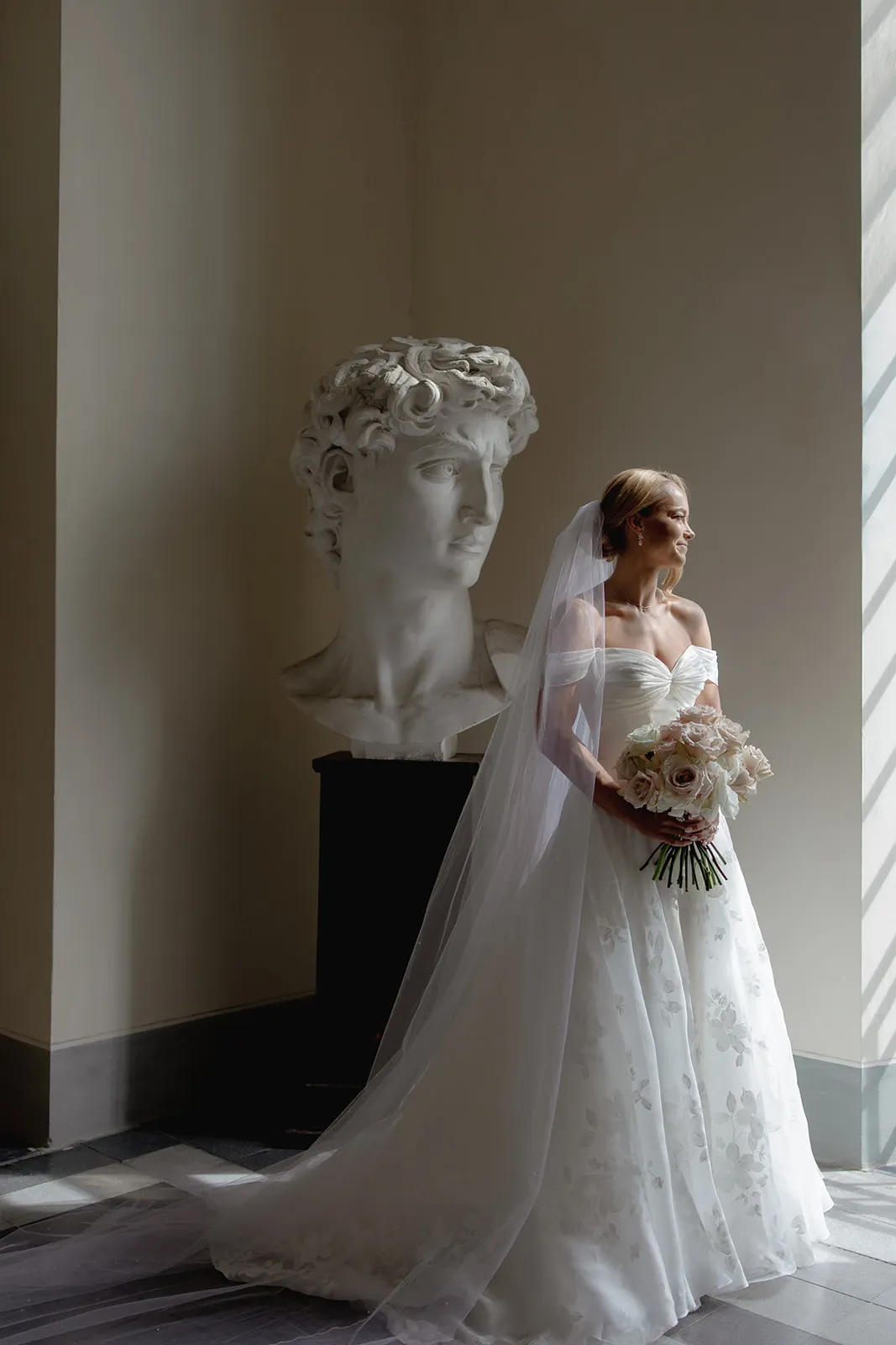 A bride in an off-the-shoulder white gown and long veil holds a bouquet of pale flowers, standing by a tall window. Behind her is a large marble bust of David, creating a classical and elegant setting with soft natural light—a perfect moment from a Destination Wedding in Italy. Italy and Tuscan Wedding Photographer