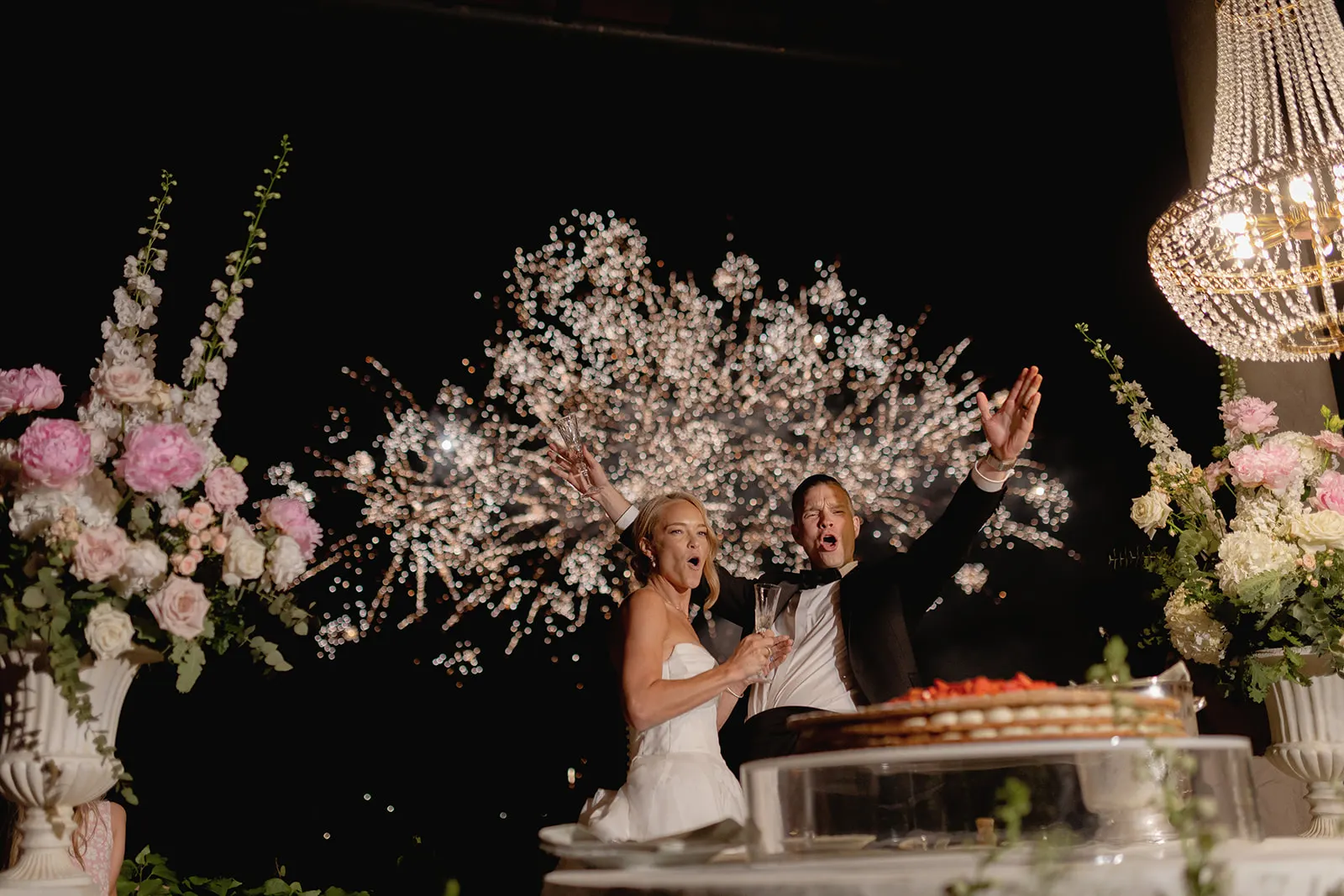 A bride and groom in wedding attire celebrate joyfully with arms raised as fireworks explode in the night sky behind them during their unforgettable Destination Wedding in Italy. Large floral arrangements and a chandelier frame the scene, while a partially visible cake sits in the foreground. Italy and Tuscan Wedding Photographer