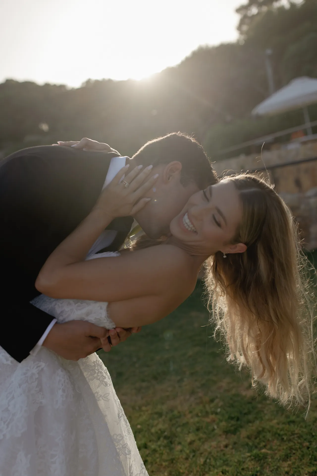 A groom in a black suit playfully dips and kisses his smiling bride in a white dress outdoors at sunset, surrounded by lush greenery and golden sunlight—the perfect romantic moment during their Destination Wedding in Italy. Italy and Tuscan Wedding Photographer