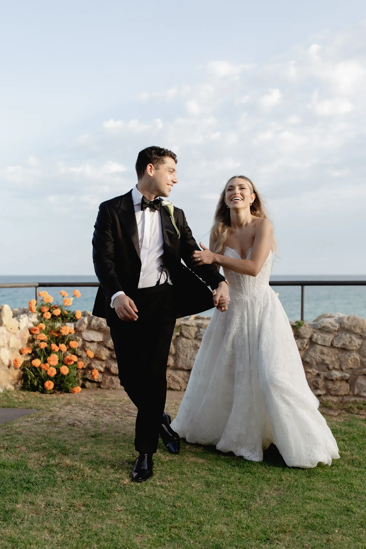 A smiling couple in wedding attire walks on grass by a stone wall with orange flowers during their Destination Wedding in Italy. The groom wears a black tuxedo, and the bride wears a white, strapless gown. They hold hands, laughing under a partly cloudy sky with the ocean in the background. Italy and Tuscan Wedding Photographer