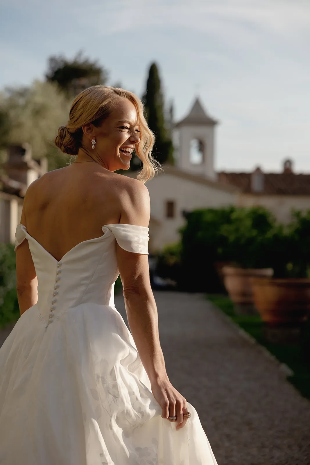 A smiling bride with light hair in a low updo wears an off-the-shoulder white wedding dress with buttons down the back. She stands outdoors on a sunlit path, holding her gown, surrounded by lush greenery and a white building with a bell tower behind her—a picture-perfect moment from her Destination Wedding in Italy. Italy and Tuscan Wedding Photographer