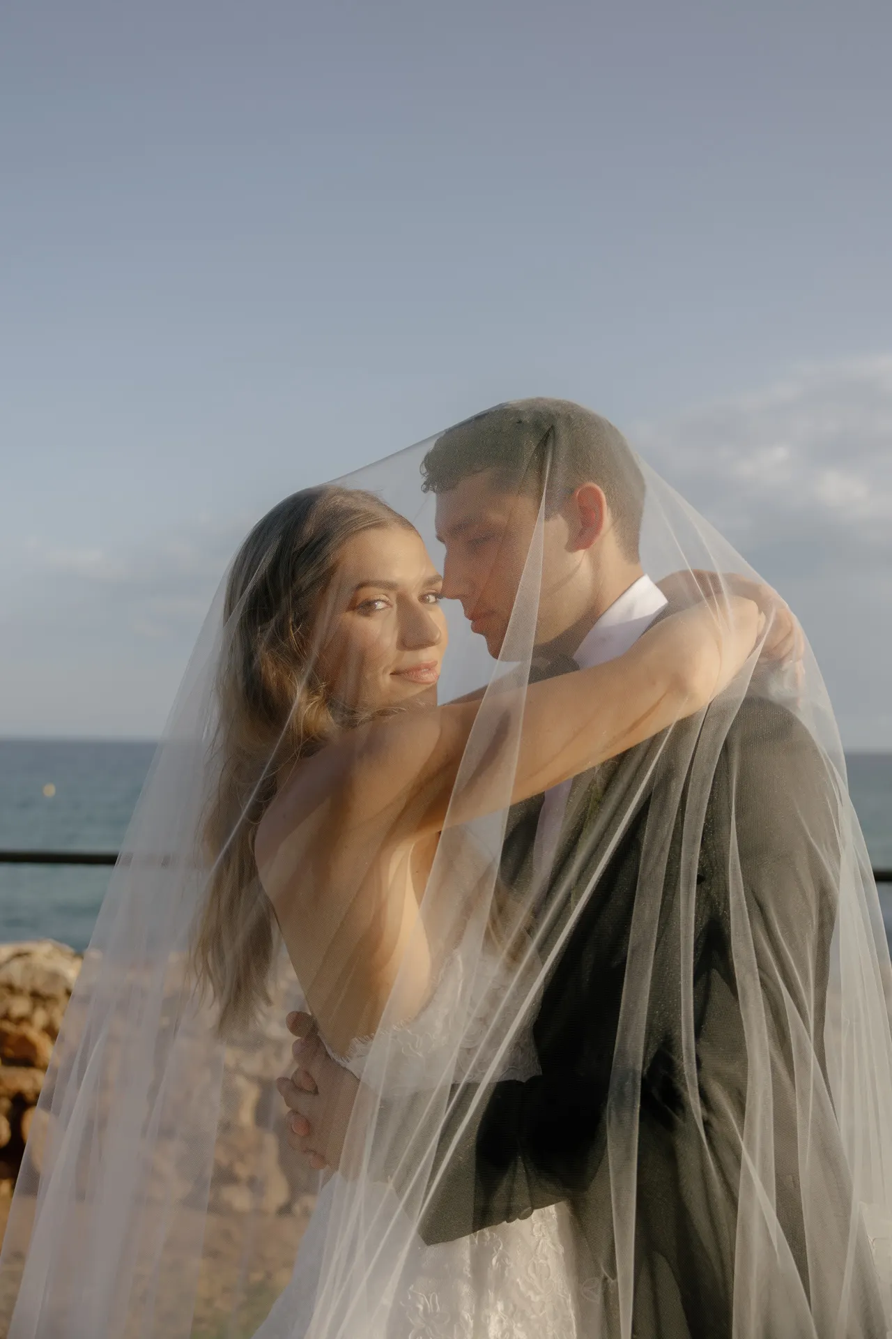 A bride and groom embrace under a sheer veil outdoors, facing the camera during their Destination Wedding in Italy. The bride looks directly at the viewer, while the groom gazes at her. The sea and a blue sky are visible in the background, enhancing the romantic atmosphere. Both appear happy and relaxed. Italy and Tuscan Wedding Photographer