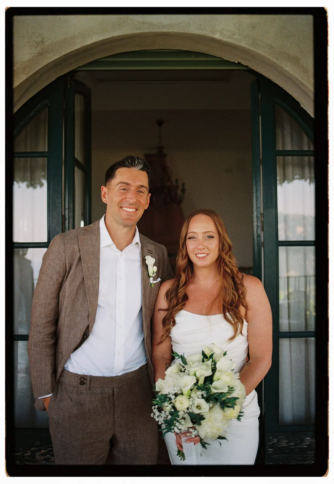 A man in a tan suit and a woman in a white wedding dress holding a bouquet stand together, smiling, in front of arched double doors with glass panes. Capturing the joy of their engagement, this moment is beautifully preserved by a Tuscany wedding photographer in a softly lit indoor space adorned with a chandelier. Italy and Tuscan Wedding Photographer