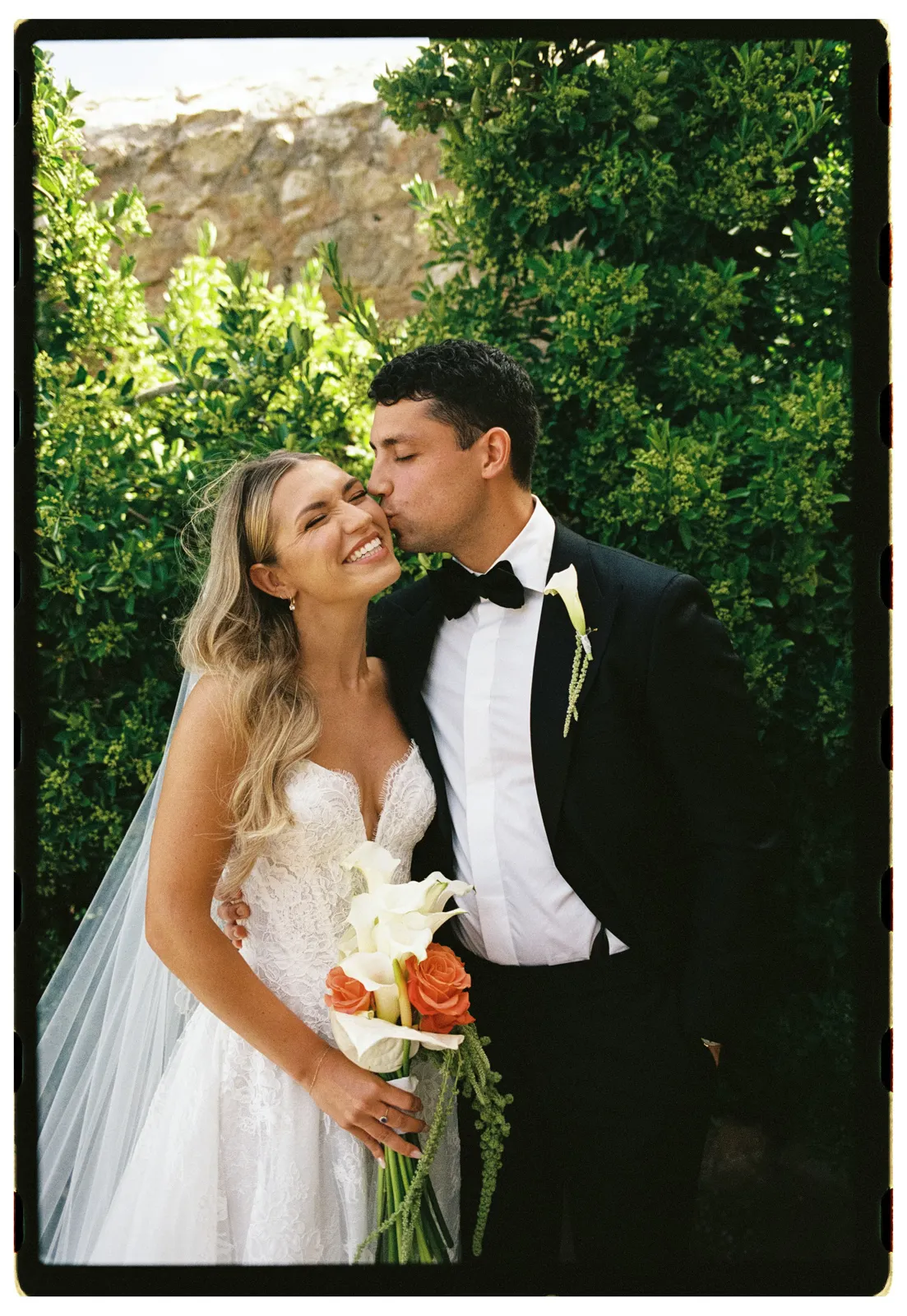 A groom in a black tuxedo kisses a smiling bride with long blonde hair in a white lace gown, holding a bouquet of calla lilies and orange roses. They stand close together outdoors, framed by lush green foliage and sunlight—an enchanting moment beautifully suited for engagement photos captured by a Tuscany wedding photographer. Italy and Tuscan Wedding Photographer