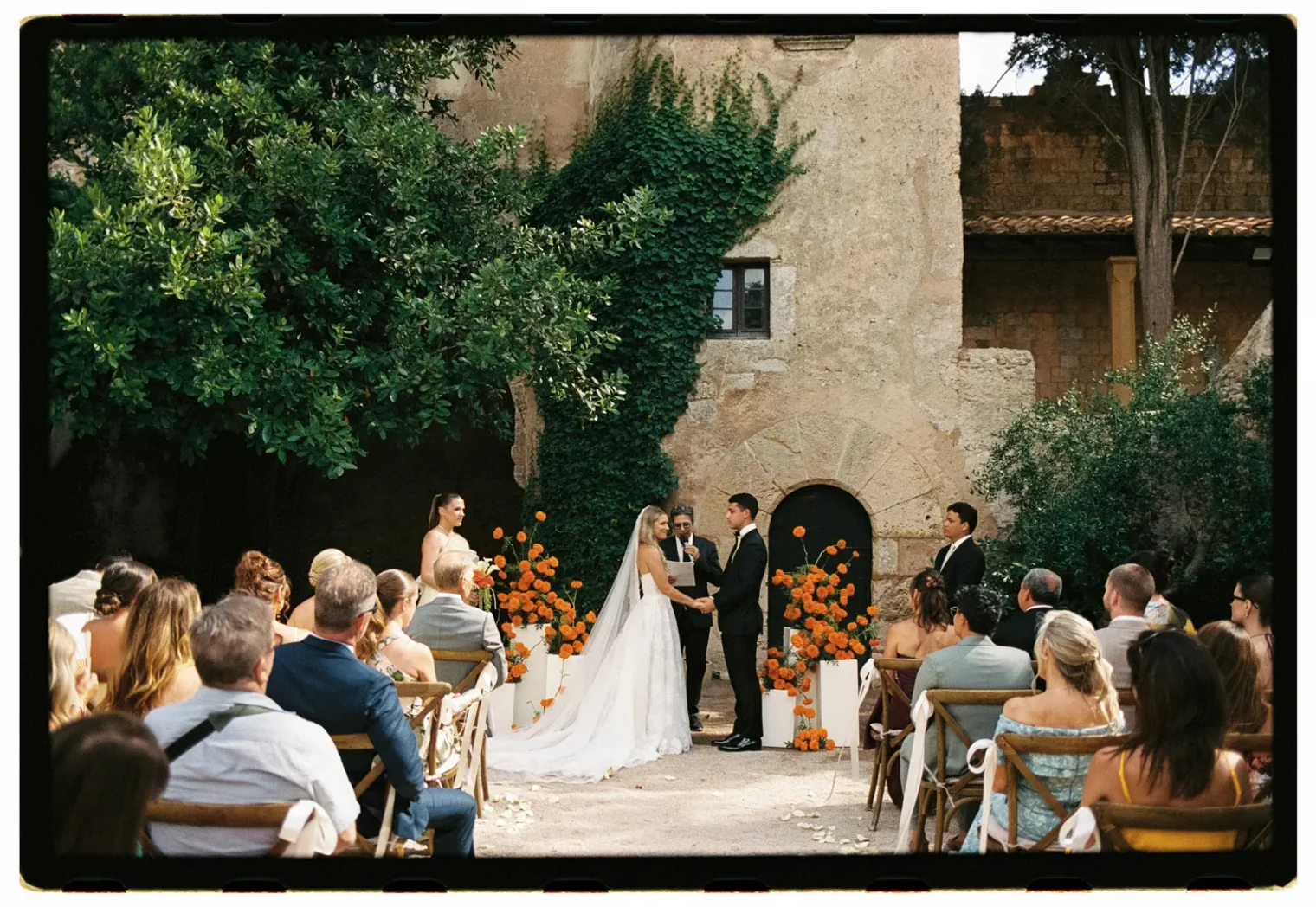 An outdoor wedding ceremony with guests seated facing a couple holding hands at the altar, beautifully captured by a Tuscany wedding photographer. The bride wears a long veil, the groom a black suit. Orange flowers decorate the aisle. Bridesmaids and groomsmen stand nearby. Ivy climbs the rustic stone building behind them, creating an enchanting backdrop reminiscent of timeless engagement photos. Italy and Tuscan Wedding Photographer