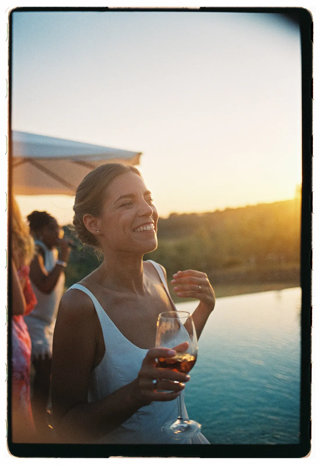 A woman in a white dress smiles and holds a glass of rosé by a pool at sunset. The background shows blurred greenery, another woman, and soft golden sunlight, creating a warm, joyful atmosphere—perfect for capturing engagement photos with the effortless elegance of a Tuscany wedding photographer. Italy and Tuscan Wedding Photographer