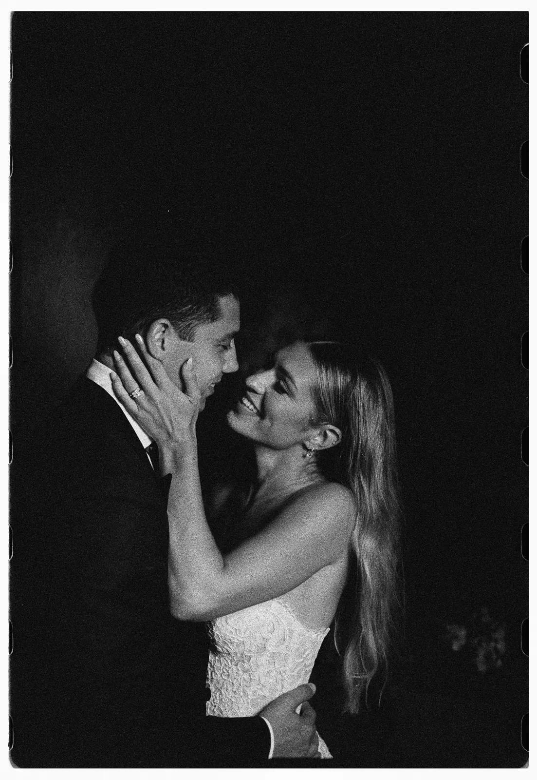 A bride in a strapless gown holds her partner’s face, smiling as they gaze into each other’s eyes in a dark, intimate setting. The groom wears a suit. Captured by a Tuscany wedding photographer, the moment feels romantic, with only their faces and upper bodies softly lit. Black and white photo. Italy and Tuscan Wedding Photographer