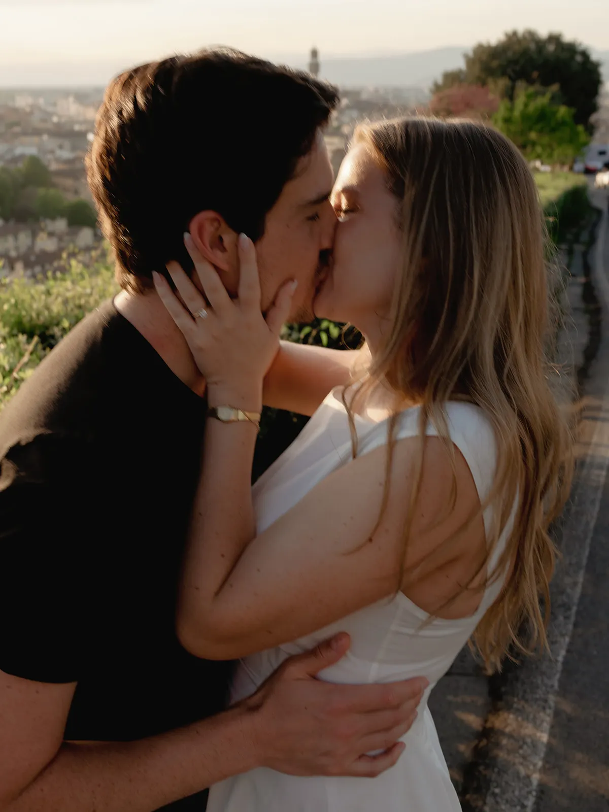 A man and woman kiss passionately outdoors at sunset in this romantic engagement photo. The woman, wearing a white dress and showing off her ring with her arm around the man’s neck, radiates happiness beside her partner in a black shirt. Greenery, a road, and a cityscape create a softly lit backdrop—perfectly capturing the magic of their special moment through the lens of a Tuscany wedding photographer. Italy and Tuscan Wedding Photographer