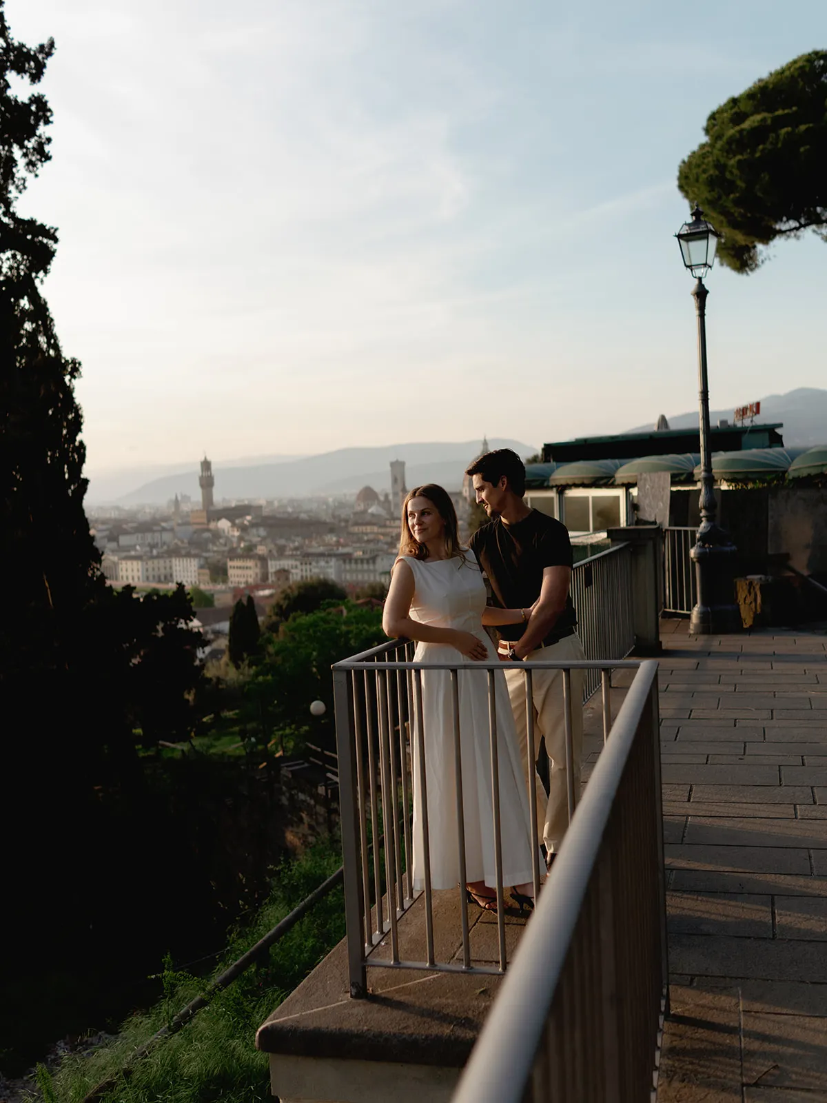 A couple stands close together on a terrace overlooking Florence at sunset, lost in the magic of their engagement. The woman wears a white dress, and the man wears a dark shirt and light pants. Captured by a Tuscany wedding photographer, the cityscape—with its historic towers and distant hills—appears bathed in warm evening light, creating timeless engagement photos. Italy and Tuscan Wedding Photographer