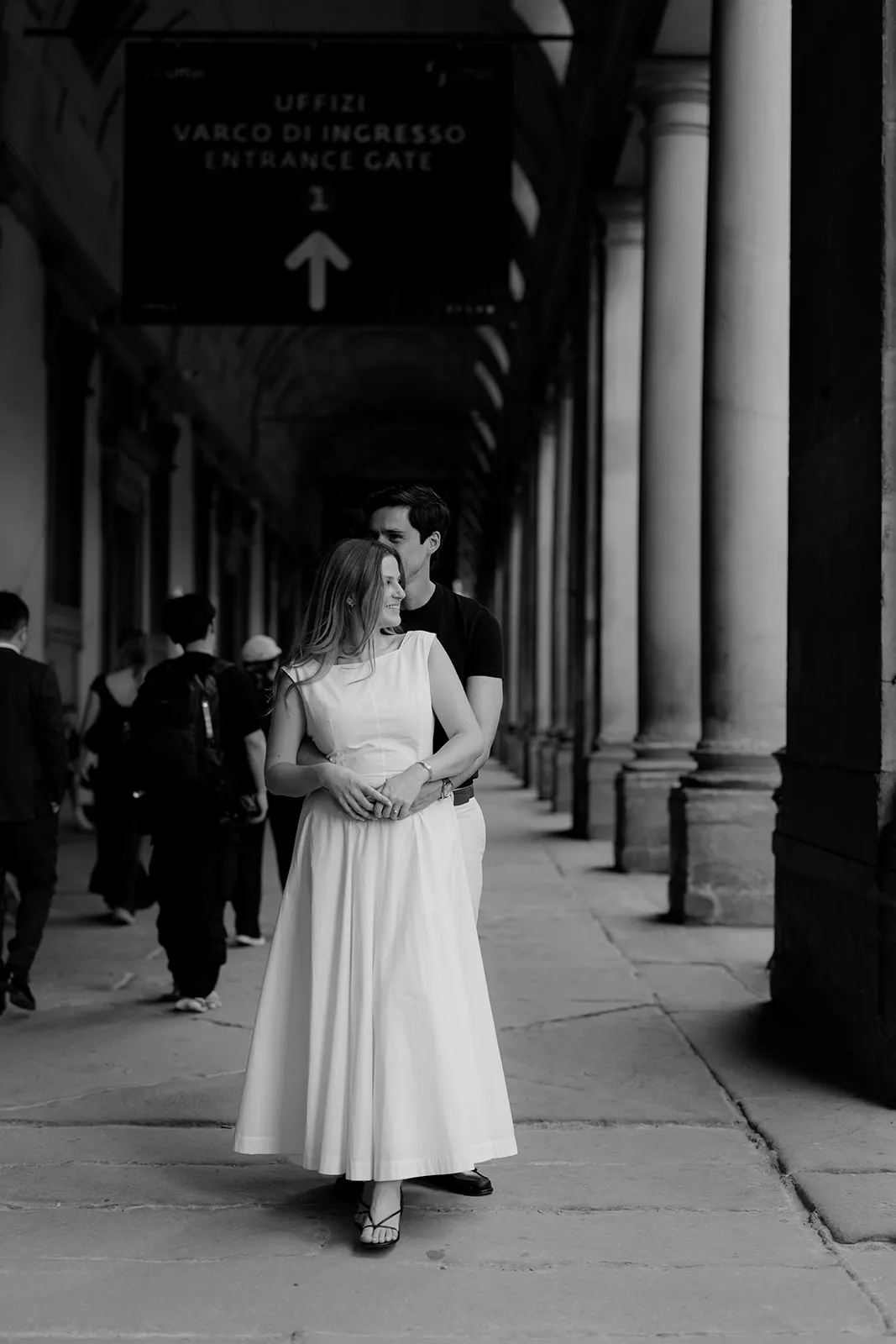 A couple stands close together in a colonnaded corridor, captured in an intimate moment by a Tuscany wedding photographer. The woman in a long white dress faces forward, while the man stands behind, arms around her waist—perfectly framing the mood of dreamy engagement photos. Other people walk in the background as a sign overhead reads Uffizi Varco di Ingresso Entrance Gate. Italy and Tuscan Wedding Photographer