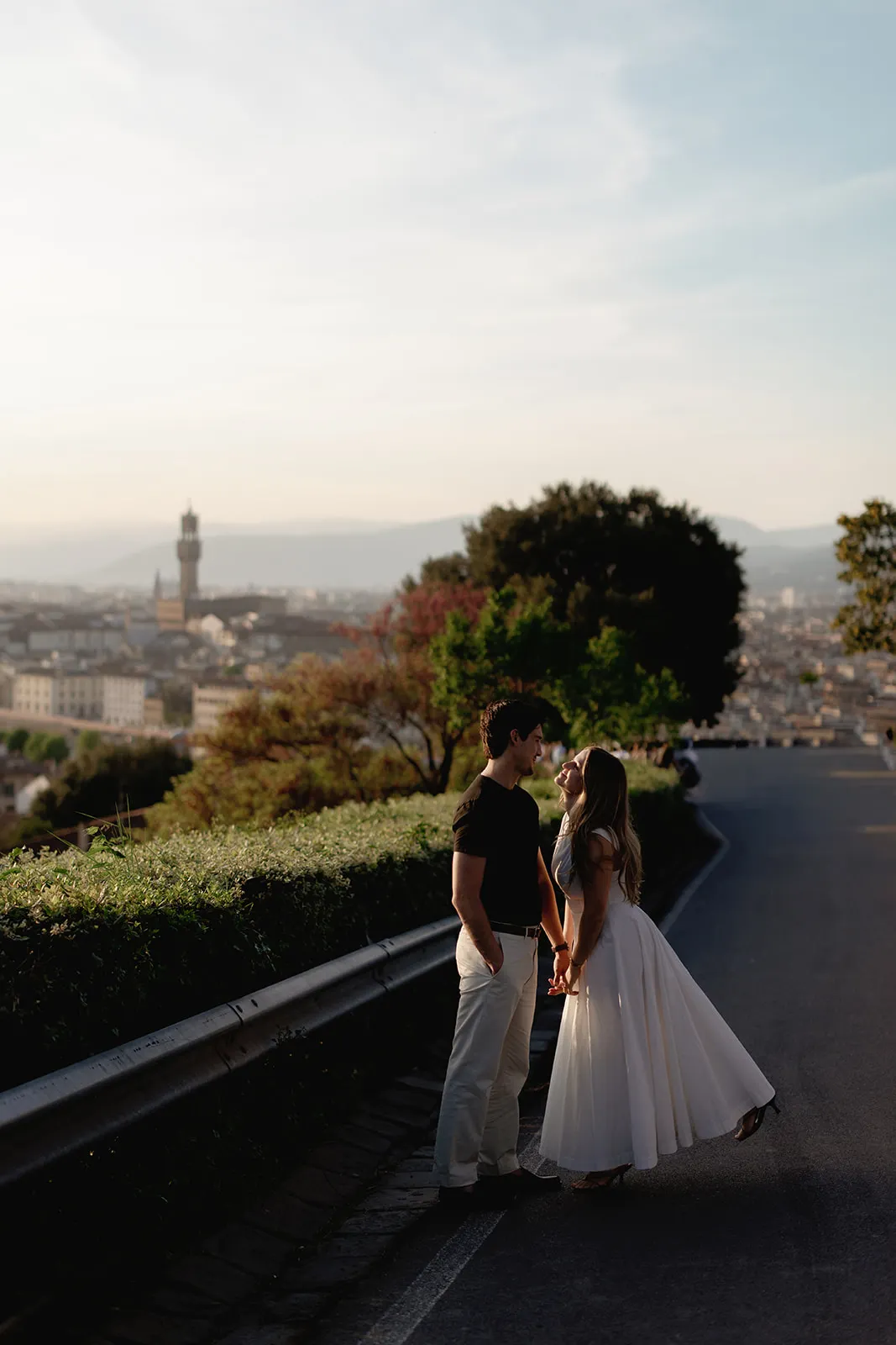 A couple stands on a scenic road, holding hands and gazing at each other in a moment of pure engagement. The woman wears a flowing white dress, the man wears light pants and a dark shirt. Florence’s cityscape and distant mountains are visible under a clear sky in the background, capturing the romantic essence sought by any Tuscany wedding photographer looking to create unforgettable engagement photos. Italy and Tuscan Wedding Photographer