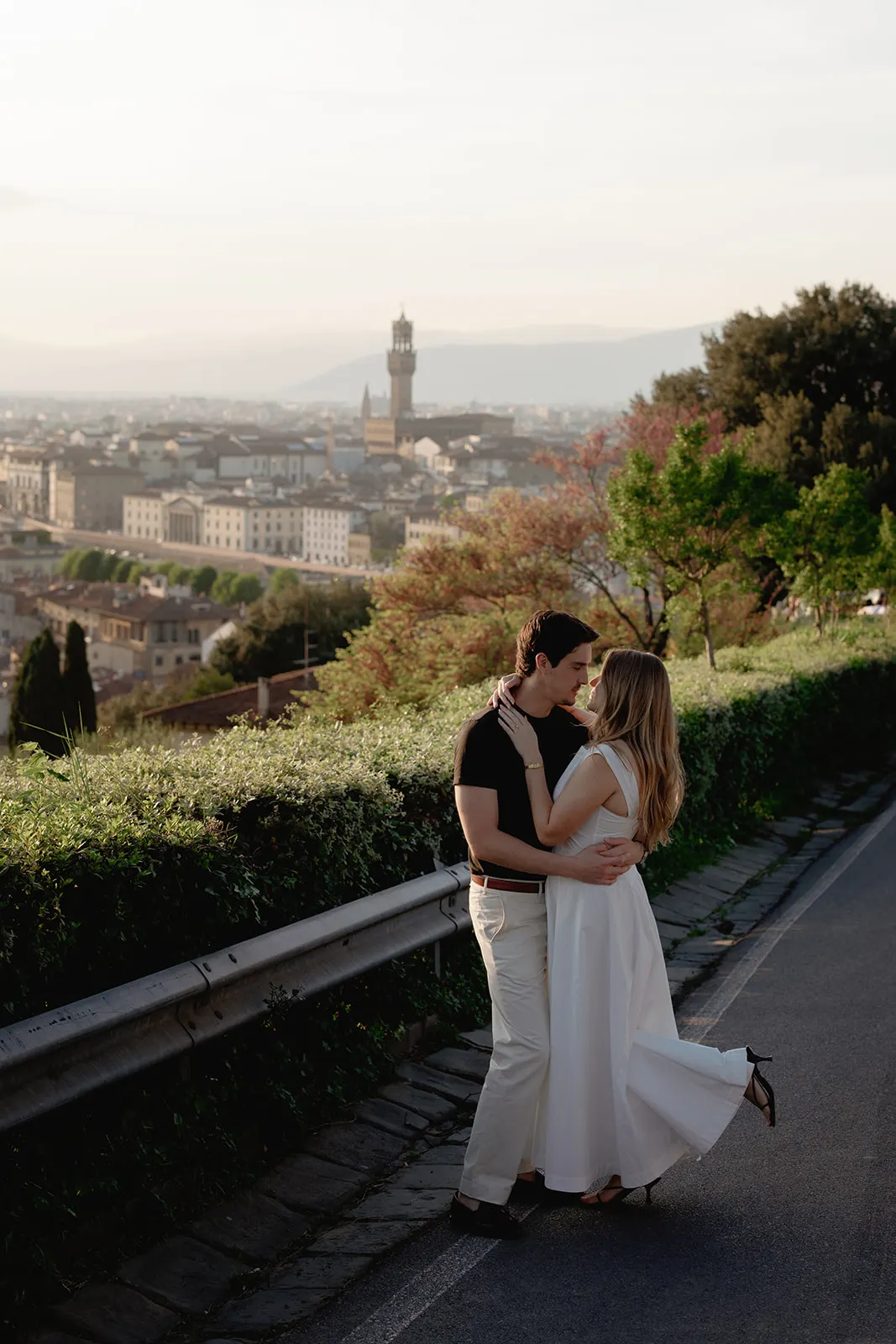 A couple embraces on a scenic road overlooking a city at sunset, perfectly capturing the magic of engagement photos. The woman wears a white dress; the man is in a black shirt and light pants. Lush greenery lines the road, with city buildings and a tall clock tower visible in the background—an idyllic setting reminiscent of moments captured by a Tuscany wedding photographer. Italy and Tuscan Wedding Photographer