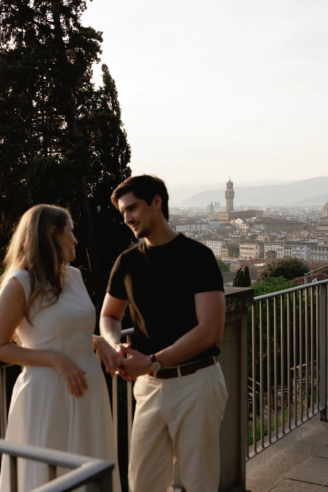 A smiling couple stands on a balcony overlooking a cityscape at sunset, capturing the magic of their engagement. The woman wears a white dress, and the man wears a black shirt and light pants. Historic buildings and a distant tower are visible in the background, surrounded by trees and soft sunlight—an idyllic moment perfect for any Tuscany wedding photographer. Italy and Tuscan Wedding Photographer