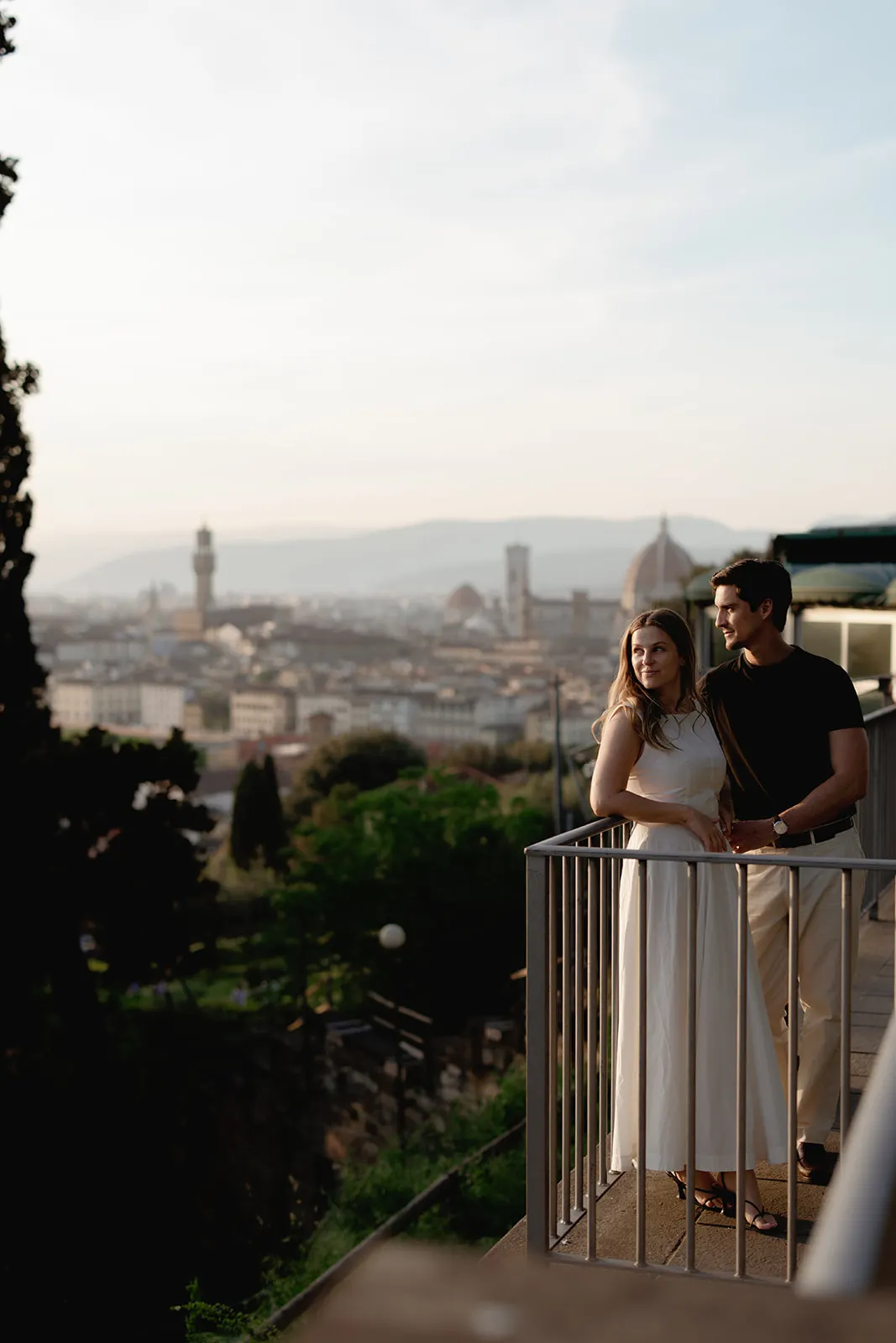 A couple stands on a balcony overlooking Florence, Italy, captured in radiant engagement photos. Both are smiling; the woman wears a white dress and the man wears a black shirt and light pants. The cityscape and distant landmarks, including the Florence Cathedral dome, are visible in the background—an unforgettable scene beautifully preserved by a Tuscany wedding photographer. Italy and Tuscan Wedding Photographer