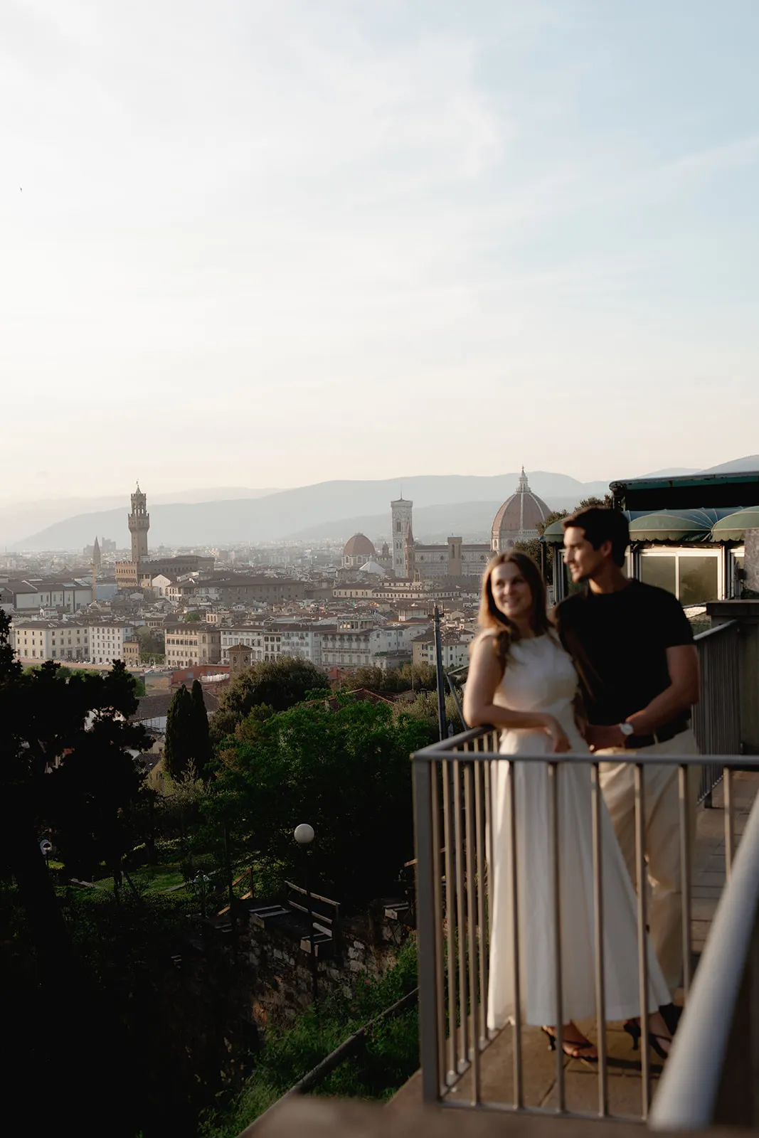 A couple stands on a balcony overlooking Florence, Italy, captured by a Tuscany wedding photographer. The woman in a white dress leans on the railing while the man stands beside her, creating a perfect moment for engagement photos. The Florence Cathedral and cityscape appear in the background under a hazy, late afternoon sky. Italy and Tuscan Wedding Photographer