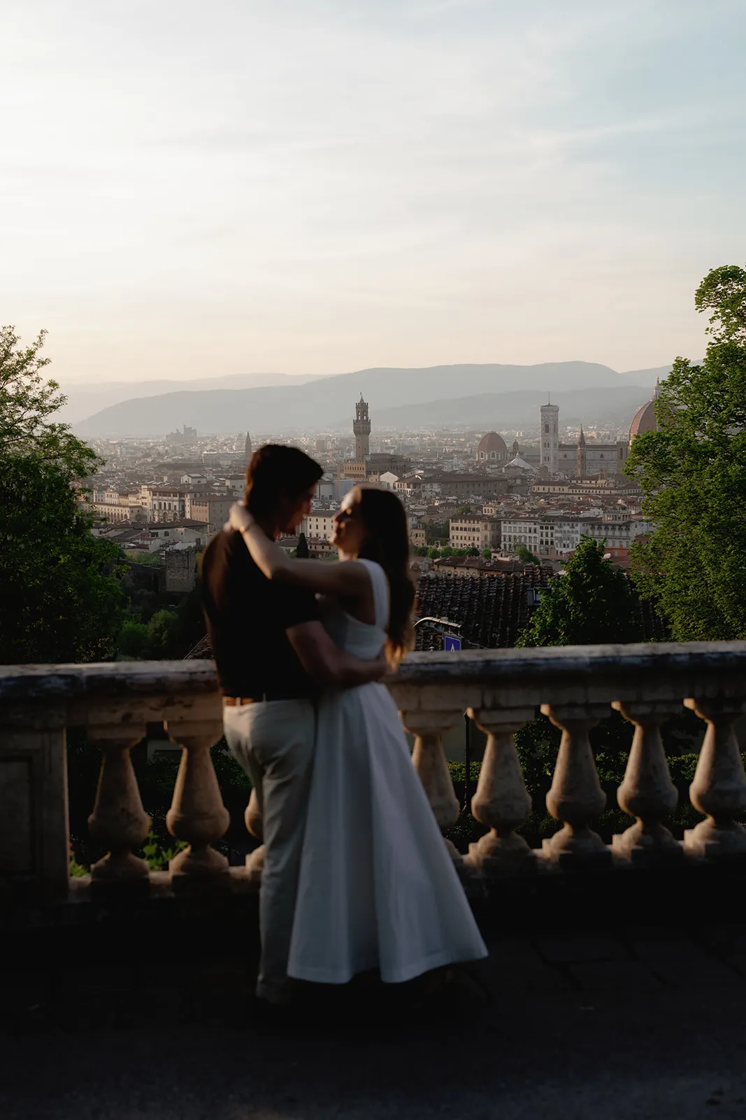 A couple embraces on a stone balcony at sunset, silhouetted against a panoramic view of Florence, Italy, with the city’s domes and towers visible in the background amid soft golden light and greenery—an unforgettable moment perfectly suited for engagement photos by a Tuscany wedding photographer. Italy and Tuscan Wedding Photographer