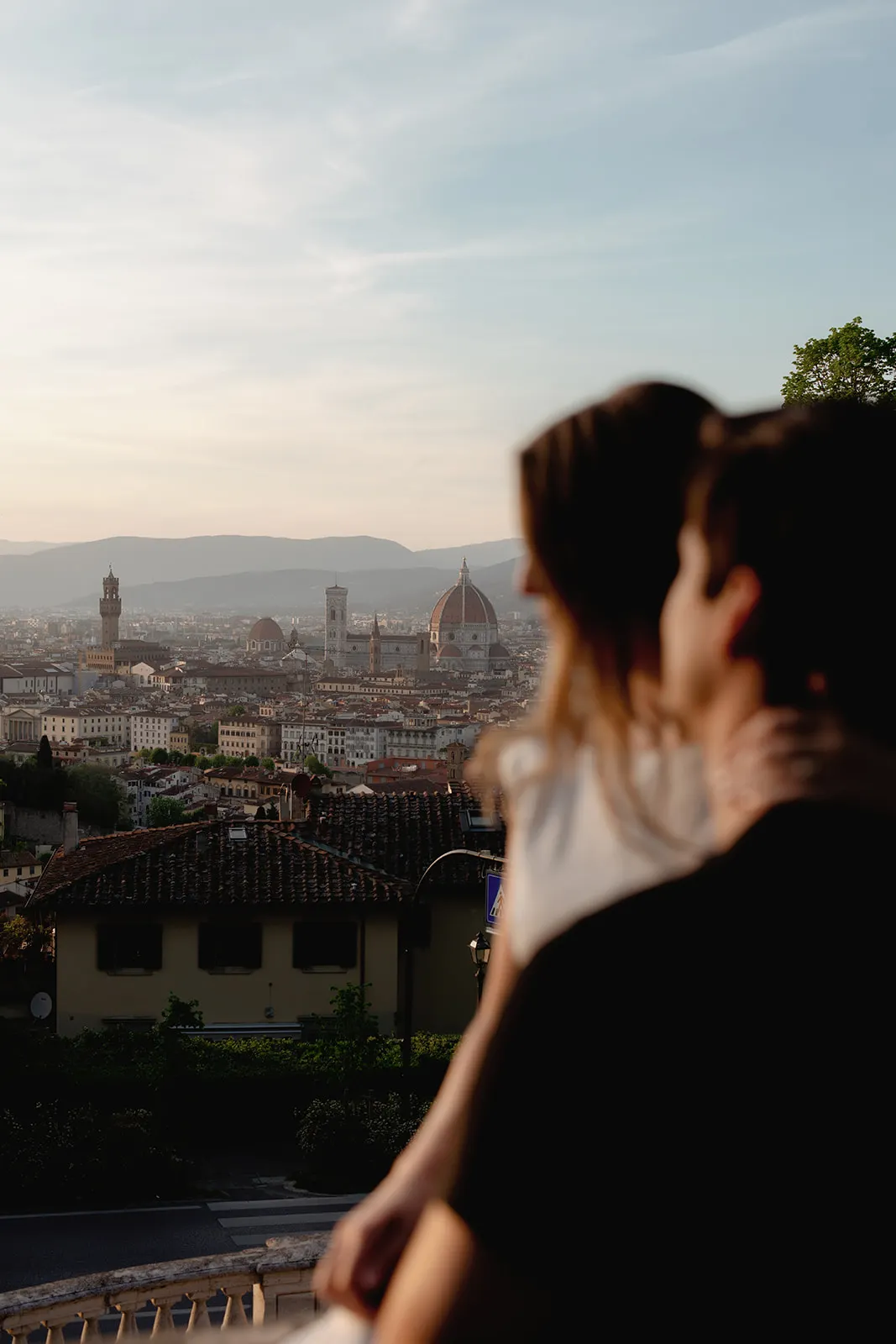 A couple in soft focus gazes over Florence at sunset, capturing a tender engagement moment. In the background, the Florence Cathedral’s dome and city landmarks are visible, set against distant hills and a pastel sky—a perfect scene for any Tuscany wedding photographer. The foreground shows rooftops and greenery, adding to the romantic atmosphere of these engagement photos. Italy and Tuscan Wedding Photographer