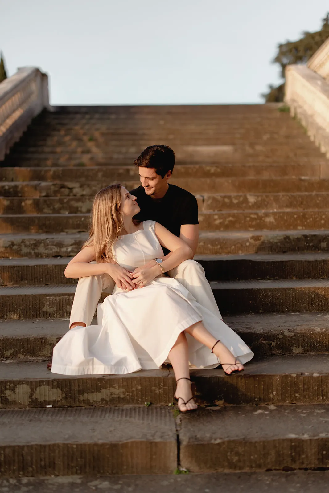 A couple sits on outdoor steps. The woman, in a white dress and sandals, leans against the man, who wears a black shirt and beige pants. They gaze at each other, smiling, with sunlight casting a warm glow on them—a perfect moment for engagement photos captured by a Tuscany wedding photographer. Italy and Tuscan Wedding Photographer