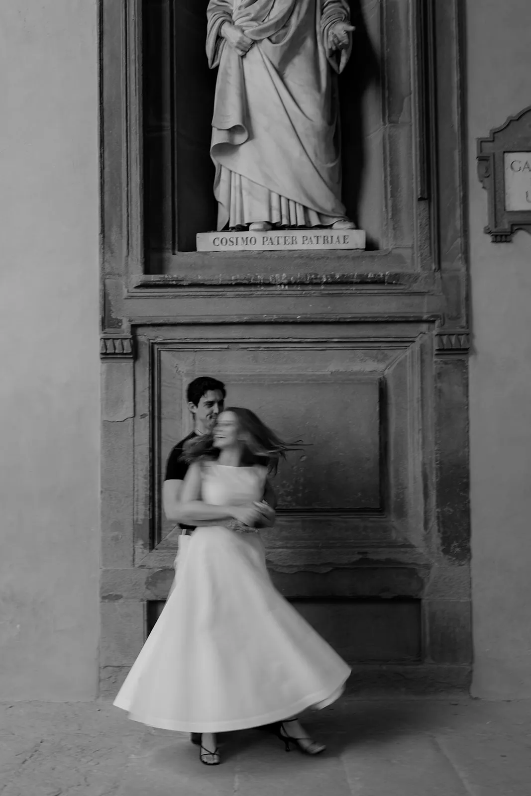 A black-and-white photo shows a man standing behind a woman in a flowing dress, holding her as she twirls. Above them is a statue labeled Cosimo Pater Patriae. The scene is set in an ornate architectural alcove with textured stone—an elegant moment perfectly captured by a Tuscany wedding photographer as part of their engagement photos collection. Italy and Tuscan Wedding Photographer