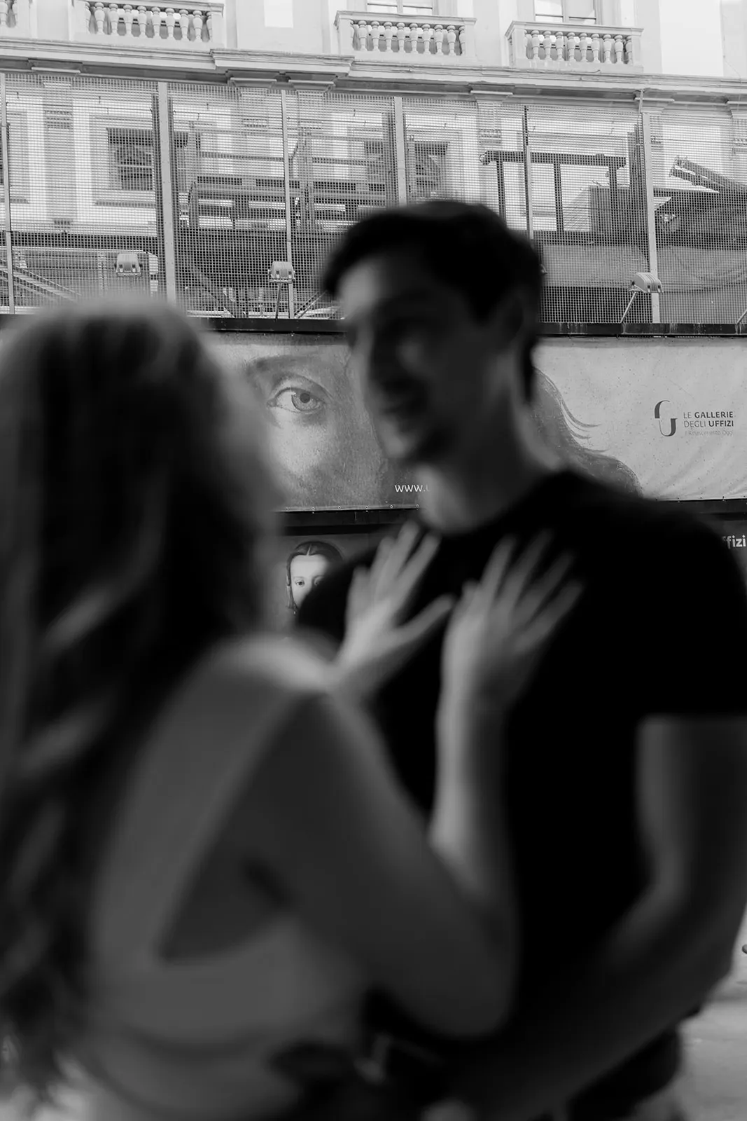 A blurred couple faces each other in the foreground, with the woman’s hands on the man’s chest, capturing an intimate engagement moment. In the background, a large photo of an eye on a building wall appears to watch them. The scene is in black and white—an evocative image that could be found in any skilled Tuscany wedding photographer’s portfolio. Italy and Tuscan Wedding Photographer