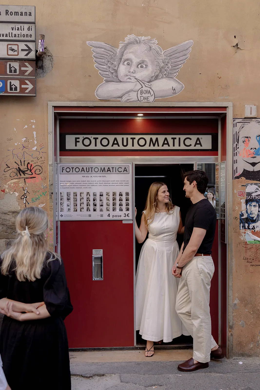 A woman in a white dress stands in a photo booth doorway, smiling at a man in a black shirt—capturing the candid joy of engagement photos. Another woman with blonde hair, wearing black, faces away. The booth is set beneath a cartoon angel mural on a tan wall covered in graffiti and street signs, reminiscent of scenes often sought by a Tuscany wedding photographer. Italy and Tuscan Wedding Photographer