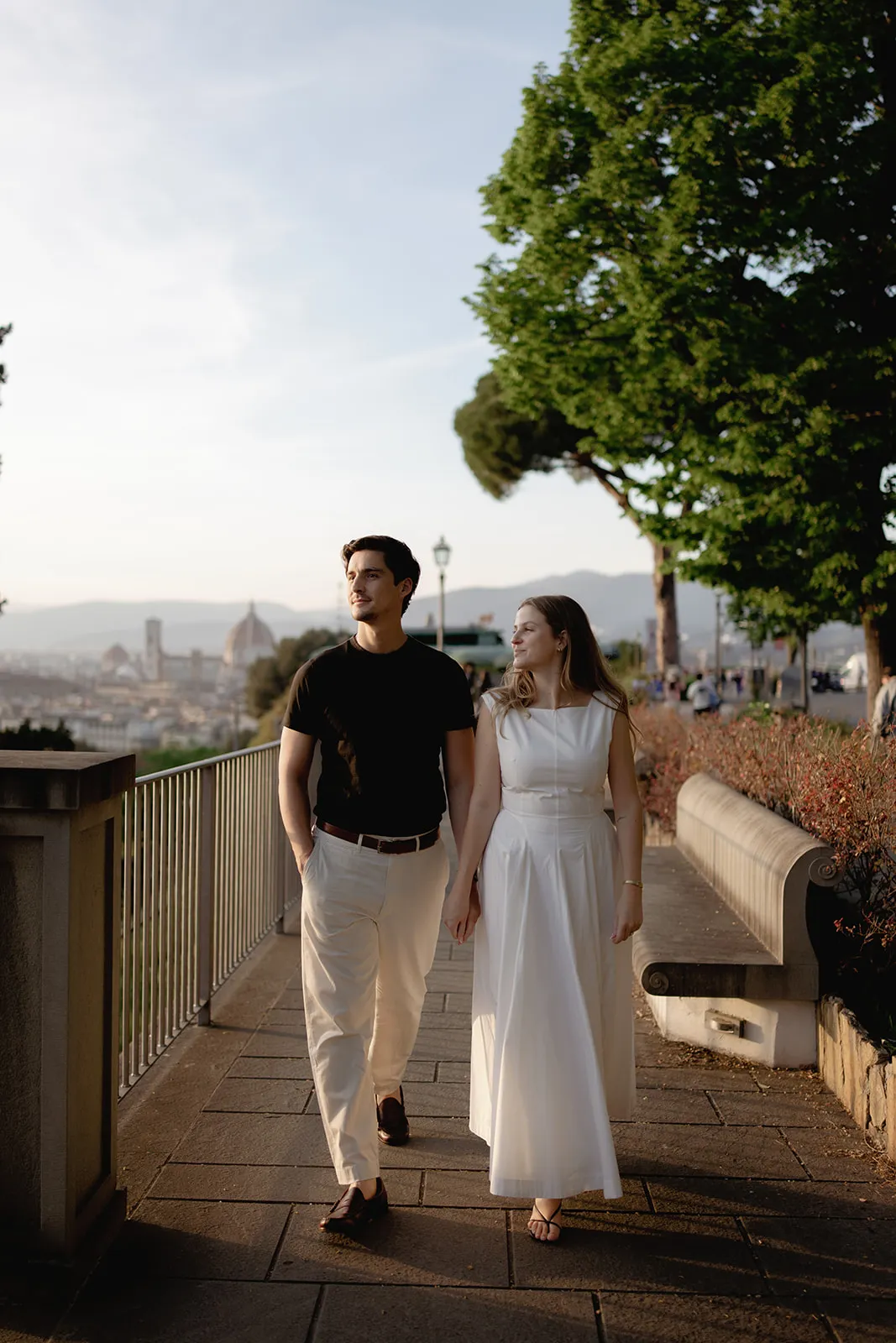 A couple walks hand in hand on a scenic path with trees and a cityscape in the background. The man wears a black shirt and light pants; the woman wears a white dress. They look at each other, smiling, with Florence’s Duomo visible in the distance—perfectly capturing the romance of engagement photos by a Tuscany wedding photographer. Italy and Tuscan Wedding Photographer