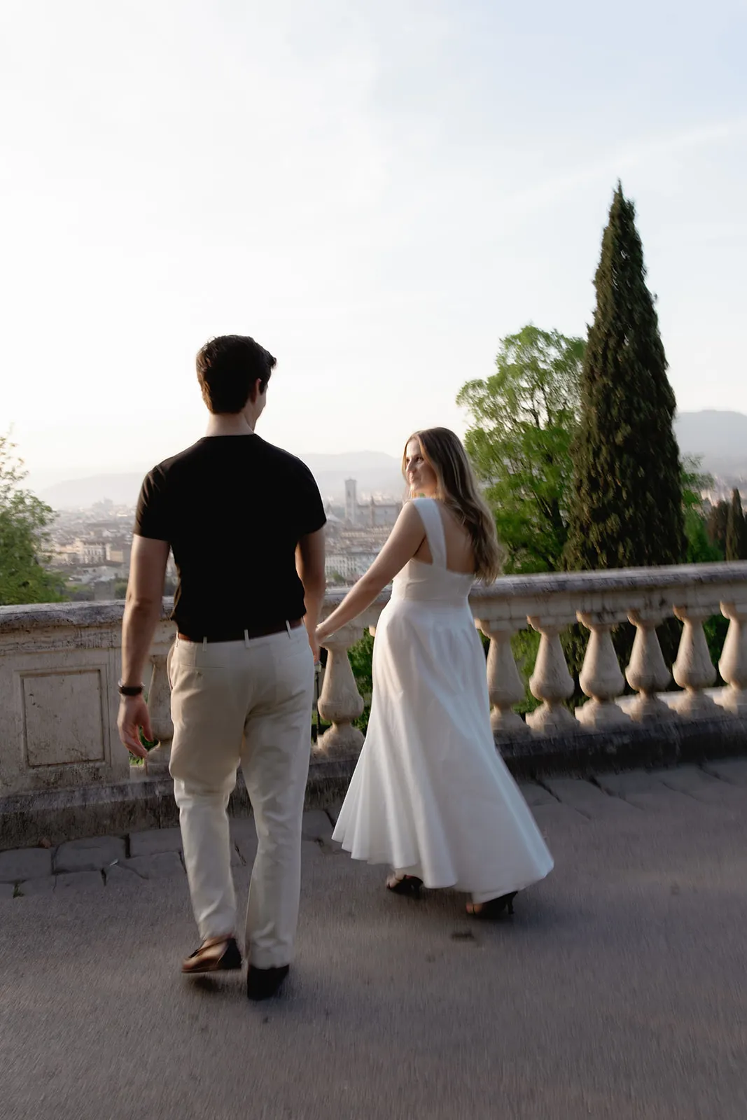 A man in a black shirt and beige pants walks beside a woman in a flowing white dress. They are outdoors on a terrace with stone railings, surrounded by trees, with a scenic city and mountain landscape in the background. The woman looks back, smiling—capturing a candid moment perfect for engagement photos by a Tuscany wedding photographer. Italy and Tuscan Wedding Photographer