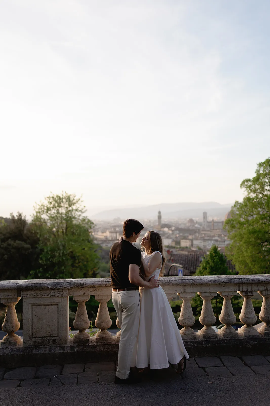A couple stands closely on a stone terrace at sunset, with the woman in a white dress and the man in a black shirt and light pants. Captured by a Tuscany wedding photographer, they gaze at each other, with a cityscape and distant hills visible in the soft, golden background. Italy and Tuscan Wedding Photographer