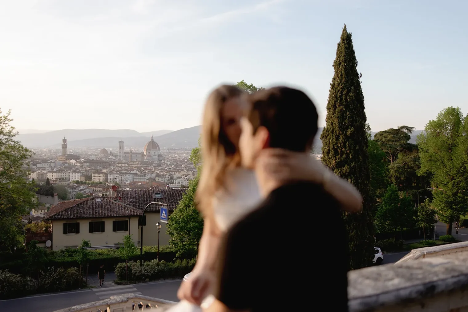A blurred couple embraces in the foreground, capturing a heartfelt engagement moment as they overlook a scenic cityscape of Florence, Italy. Perfect for engagement photos by a talented Tuscany wedding photographer, the Duomo and historic buildings are visible in the distance, framed by green trees and a tall cypress, with mountains in the hazy background under a clear sky. Italy and Tuscan Wedding Photographer