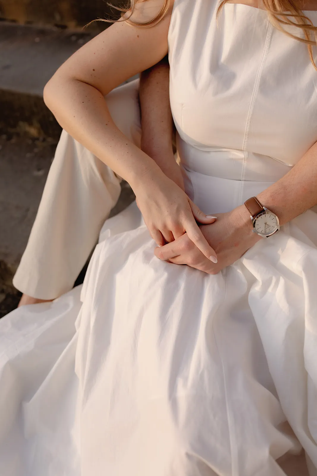 A woman wearing a sleeveless white dress sits outdoors, possibly during engagement photos. Her hands rest gently in her lap, and she wears a watch with a brown leather strap on her left wrist. Only her arms, torso, and part of her face and hair are visible. Italy and Tuscan Wedding Photographer
